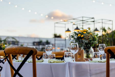 Romantically set dinner table with wine glasses and floral centerpiece, perfect for a wedding, elegant party, or outdoor evening celebration. Warm lights and soft bokeh create an intimate and luxurious atmosphere.の写真素材