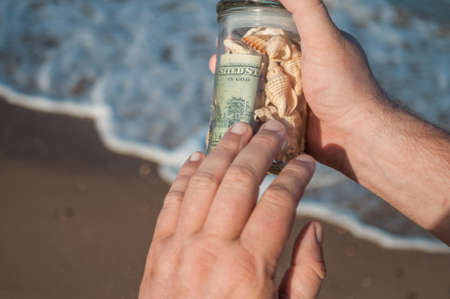 male hands holding a glass jar with money and shells found on the shore of the seaの写真素材