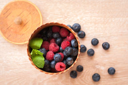 juicy raspberries and blueberries in a wooden bowl and a few scattered on the tableの写真素材