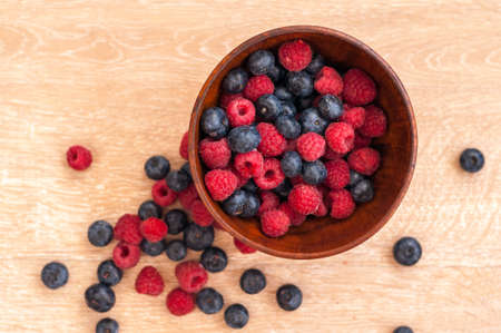 juicy raspberries and blueberries in a wooden bowl and a few scattered on the tableの写真素材