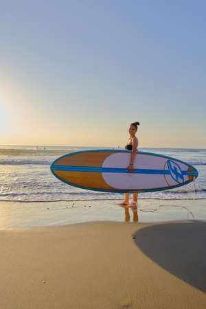 young beautiful girl in a bathing suit with a surfboard at dawnの写真素材