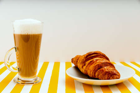 Coffee with milk in an Irish glass next to a croissant on a white saucer on a striped white and yellow table. Close-up on a white background.の写真素材