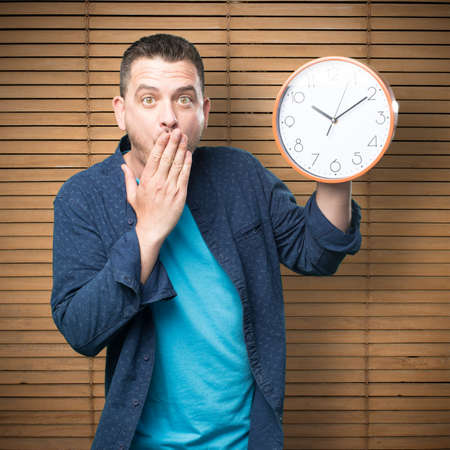 Young man wearing a blue outfit. Holding a clock. Looking surprised.