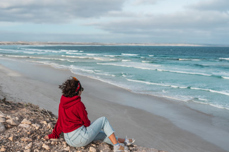 Women with short hair and red hoodie sitting in the rocks closed to the beach and looking to the sea