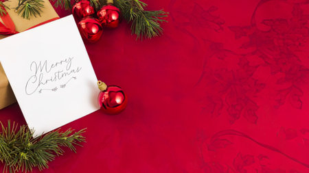 a white christmas card with 'merry christmas' written on it lies on a red velvet surface surrounded by green pine branches red baubles and a gold gift box