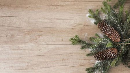 The image features a close-up view of two pine cones surrounded by green pine needles on a light-colored wooden surface. The pine cones are brown and have a textured surface. The pine needles are dusted with snow or frost, adding a touch of winter wonder to the scene. The wooden surface appears to be made of light-colored wood planks, possibly a table or floor.