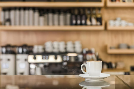 a white coffee cup on a saucer sitting on a shiny table in a blurred cafe background with shelves and coffee machines