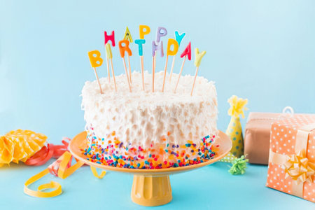 a white frosted birthday cake with colorful sprinkles on the bottom and a happy birthday sign on top sits on a yellow cake stand on a blue table with party supplies and gifts