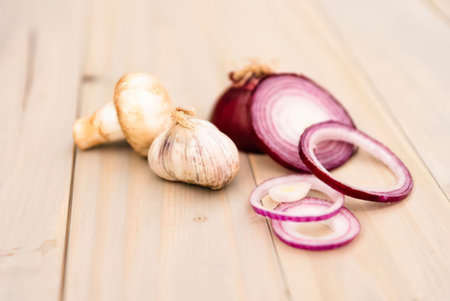 a sliced red onion and two garlic bulbs are placed on a light-colored wooden surface. the red onion is sliced into thin rings, with one ring standing up. the garlic bulbs are whole and unpeeled.