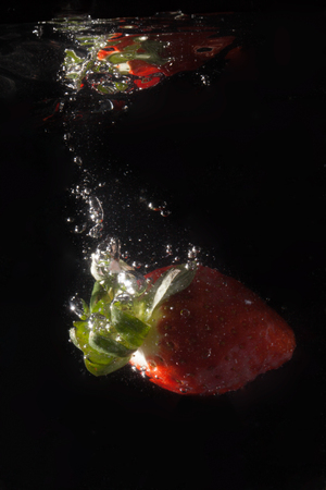 A fresh strawberry splashing in water against a black backdrop clearly showing the bubbles of air in the water and a reflection of the fruit against the surface.の写真素材