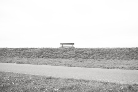 A single empty wooden bench on the horizon above a grass field against a large grey clouded sky depicting loneliness, grieve, being alone, sadness in black and white or monochromeの写真素材