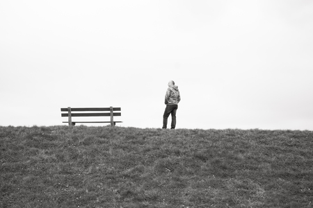 A single person standing next to a single empty wooden bench on the horizon above a grass field...
