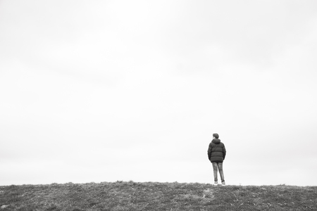A single person standing on the horizon above a grass field against a large grey clouded sky depicting loneliness, thinking, pondering, grieve, being alone, sadnessの写真素材
