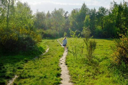 Unrecognizable young girl with dog walking on the road in forest with her back to camera.