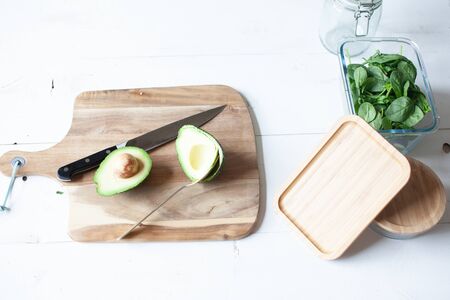 Wooden cutting board with spinach and avocado and a knife and glass jars on white wooden background