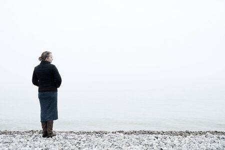 Woman wearing warm clothes and a purse looking out over a foggy sea from a pebble beach on the shoreの写真素材