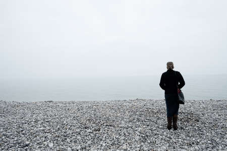 Woman wearing warm clothes and a purse looking out over a foggy sea from a pebble beach on the shore