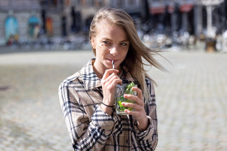 Outdoor lifestyle close up portrait of attractive lovely young woman with long hair holding and drinking a cocktail drink against a city background. High quality photoの写真素材