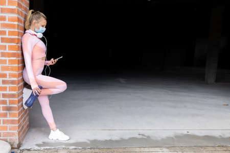 Fit pretty young woman using smartphone and headphone while resting after exercising, leaning...