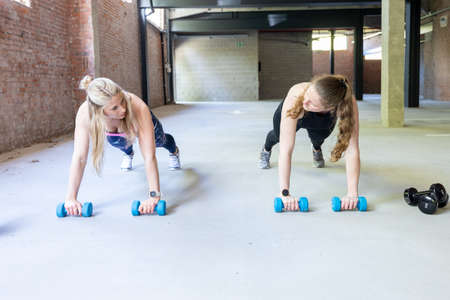 Two young attractive women doing push ups inside with barbells. Fitness woman working out in a cross...