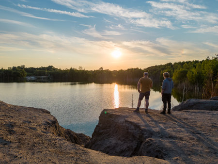 Aerial drone shot of the silhouettes of two people father and son watching the dramatic and colorful sunset with orange, yellow and grey clouds over the trees on the shore of peaceful forest lake . High quality photoの写真素材