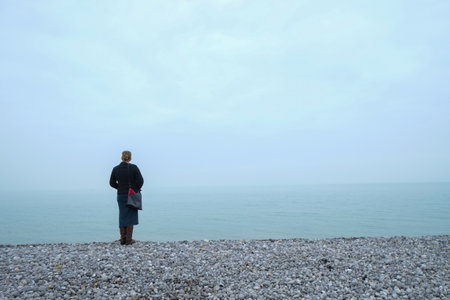 Woman wearing warm clothes and a purse looking out over a foggy sea from a pebble beach on the shoreの写真素材
