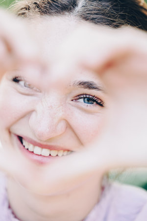 Close up image of smiling woman out in nature making a heart shape with hands. Pretty joyful caucasian woman laughing at camera outside. Healthy lifestyle, self love and body care concept. High quality photoの写真素材