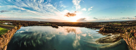 An immersive panoramic aerial view of a dramatic and vibrant sunset painting the sky over a serene forest lake. The play of colors creates a stunning spectacle, reflecting the suns setting hues on the calm lake surface. Panoramic Aerial View of a Dramatic and Colorful Sunset Over a Forest Lake. High quality photoの写真素材