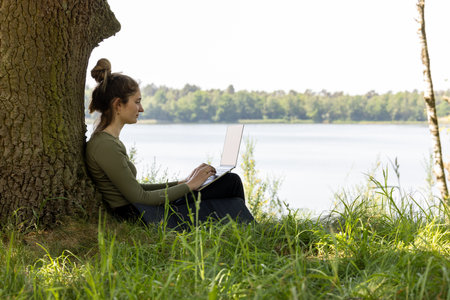 A young and talented female freelancer finds herself in a blissful paradise, working with her laptop while surrounded by the breathtaking view of a serene forest and a tranquil lake. This idyllic setting grants her the freedom of teleworking, as she enjoys the distance from the usual confines of a traditional office, allowing her creativity to flow amidst the beauty of nature. Laptop with a View: Female Freelancer Embracing Natures Freedom. High quality photoの写真素材