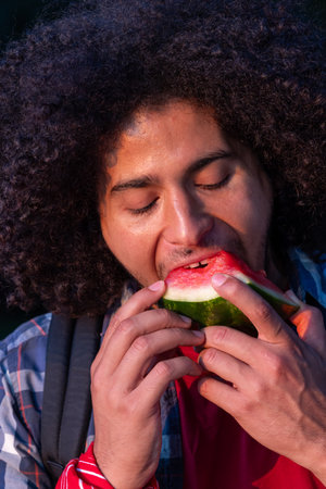 A heartwarming close-up captures the pure joy of a handsome young brown man as he enthusiastically savors a delicious slice of watermelon. His infectious happiness shines through as he relishes each bite, his eyes gleaming with delight. This candid moment showcases the simple pleasure of indulging in a summertime treat with unbridled enthusiasm. Close up of happy handsome young brown man very enthusiastically eating watermelon sliceの写真素材