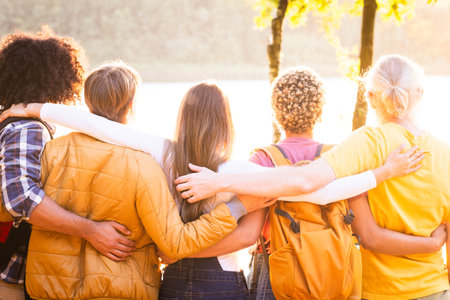 Friendship, multi ethnic group, togetherness, traveling, vacation, holidays, togetherness, sightseeing. Young people with backpacks standing close hugging admiring the natural sunset view over the forest lake. High quality photoの写真素材
