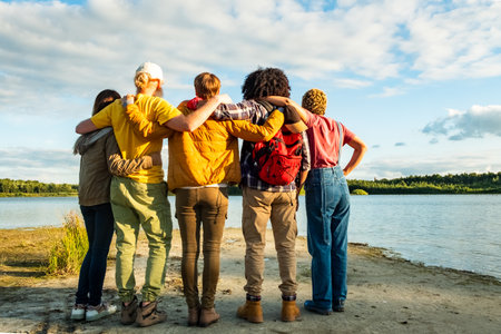 Wanderlust lifestyle, Back view of group of young multi racial friends camping standing hugging on shoulder together on the shore of a forest lake. High quality photoの写真素材