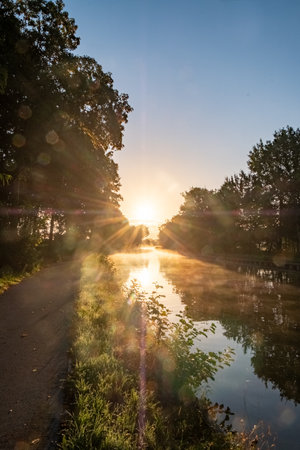 Experience the breathtaking beauty of a radiant sunrise as it casts its golden glow over a serene canal nestled between enchanting trees. The tranquil water reflects the suns brilliance, creating a mesmerizing scene that captures the peaceful magic of natures morning spectacle.Radiant Sunrise: Sunlit Canal Reflecting Amidst Tranquil Trees. High quality photoの写真素材