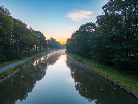 Embrace the tranquility of nature with this enchanting low-angle view capturing a picturesque canal gracefully winding its way between a majestic canopy of towering trees. Underneath a soothingly clouded sky, the serene waters reflect the lush greenery, creating a scene that invites contemplation and peace. Canal Serenity: Tranquil Waters Amidst Towering Trees. High quality photoの写真素材