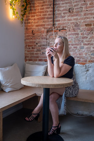 Young blonde millennial woman in a stylish blouse and skirt sits at a cafe table. Holding her coffee with both hands near her face, she gazes outside with a radiant smile, lost in pleasant thoughts. Blonde Millennial Woman Enjoying Coffee in Cafe. High quality photoの写真素材