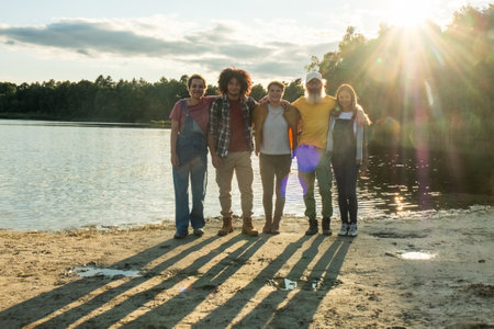 A diverse group of millennial friends stands on the forest lake shore, walking toward the camera in the warm glow of the setting sun, casting long shadows on the sandy beach. Multiracial Friends Walking on Lake Shore. High quality photoの写真素材
