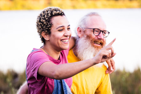 This high-quality photo beautifully captures a unique moment of connection between a young South American millennial woman and an older, grey-bearded Caucasian man. Together, they walk closely and joyfully in nature, exemplifying fun and ageless companionship. Ageless Joy: Diverse Couples Nature Walk. High quality photoの写真素材