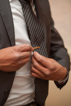 In this image, a mans hands are captured in the act of fastening a silver tie clip, adding the final...