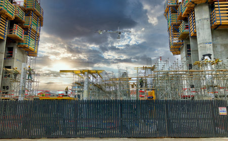 This dynamic scene captures the hustle of an urban construction site as the day winds down. Towering cranes and scaffolding outline the emerging skeleton of new buildings against a dramatic sky. The sun peeks through the clouds, casting a radiant glow and elongating shadows across the site, while the surrounding fencing marks the boundary between development and the existing cityscape.の写真素材