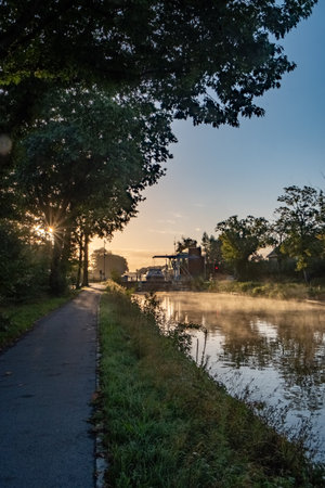 In this serene scene, the morning sun peeks through the trees along a tranquil waterway, casting a brilliant reflection on the surface of the water. A path runs alongside the river, inviting a peaceful morning walk. Mist rises from the water, adding a touch of mystique to the dawn atmosphere, while a boat sits moored in the background, awaiting the days journey. Riverside Repose: Sunrise by the Waterway. High quality photoの写真素材