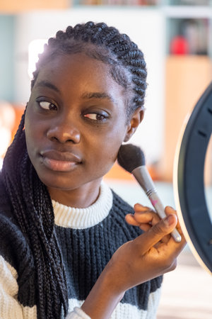 This portrait captures a woman in the midst of applying makeup, brush in hand, with a thoughtful expression. Her focus is directed towards the mirror, reflecting a moment of self-care and preparation. The soft lighting from the ring light softly accentuates her features, highlighting the personal and intimate nature of the routine. The womans braided hair and the casual attire suggest a relaxed yet attentive approach to her appearance, embodying a blend of confidence and introspection. Makeup Moment of Reflection. High quality photoの写真素材