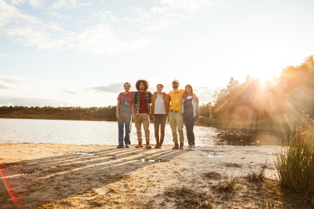 The image captures a relaxed and content group of friends standing together by a lake, with the sun setting behind them. The group is diverse, including a young Caucasian boy, a Middle-Eastern man, a Hispanic woman, and an elderly Caucasian man. The soft evening light washes over them, creating a serene atmosphere and casting long shadows on the ground. The open space and natural setting evoke feelings of peace and companionship. Lakeside Unity: A Groups Golden Hour Gathering. High quality photoの写真素材