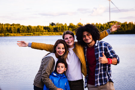 The image depicts a joyful group of four individuals in a picturesque lakeside setting during dusk. The group consists of a young Hispanic woman with long, straight hair, a Caucasian boy in a blue jacket exuding youthful enthusiasm, a Caucasian teenage boy with a bright smile, and a Middle-Eastern young man with curly hair giving a thumbs up. They are all smiling broadly, arms around each other, capturing a moment of carefree happiness. The lake behind them reflects the calming tones of the evening sky, enhancing the feeling of a relaxed and enjoyable outing. Lakeside Bonding: A Group Portrait at Dusk. High quality photoの写真素材