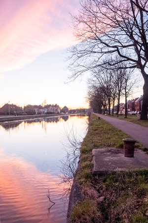 This image offers a portrait-style view of a serene canal during the soft light of the evening. The sky, painted in strokes of pink and lilac, is perfectly reflected in the canals still water, creating a symmetrical harmony. The tree line forms a natural border that guides the eye along the waterway, while the path invites a walk in the quiet of the approaching night. This composition evokes a sense of peaceful living alongside nature, with the residential backdrop adding a layer of human presence to the natural tranquility. Evening Harmony: Canal-side Repose. High quality photoの写真素材