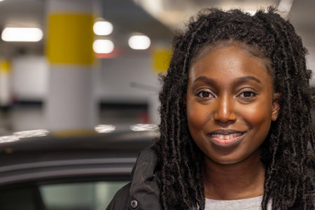 This is a warm, engaging portrait of a young woman smiling directly at the camera. She appears to be standing in a parking garage, indicated by the cars and the structural elements in the soft-focused background. Her relaxed, confident demeanor contrasts with the typically impersonal nature of such a setting, adding a humanizing element to the urban landscape. The lighting in the garage casts a soft glow on her face, highlighting her features and the texture of her hair. Urban Confidence: Portrait of a Smiling Young Woman. High quality photoの写真素材