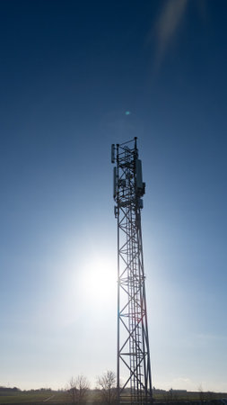 This vertical image features a towering silhouette of a telecommunication tower set against the backdrop of a clear blue sky, with the sun shining brightly and creating a flare effect. The tower stands tall, its intricate metal structure outlined by the backlight of the sun, which seems to crown the top of the tower with rays of light. The sun flare adds a dynamic element to the image, suggesting the towers role in harnessing and distributing the invisible waves of communication that connect us all. Solar Flare: Telecommunication Tower Against the Sun. High quality photoの写真素材