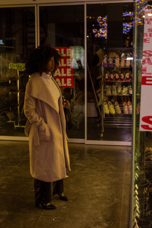 The warmth of a city evening is encapsulated in this image, where a woman stands by a shop window,...