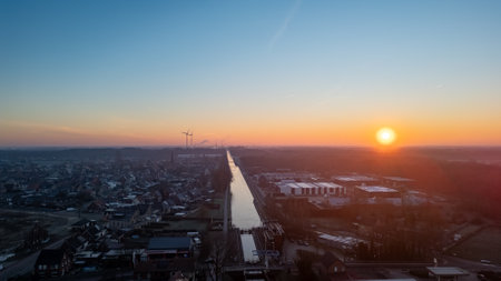 This image captures the serene moment of sunrise, seen from an elevated viewpoint over an urban landscape. The rising sun bathes the scene in a soft, golden light, contrasting with the cool blues of the dawn sky. A straight road cuts through the urban area, leading the eye toward the horizon where the silhouettes of wind turbines are visible, symbolizing the blend of urbanization with sustainable energy solutions. The calmness of the morning is palpable, with the city still in slumber as the new days light gently awakens it. The photograph encapsulates the hope and tranquility of early morning in the city, with the promise of a new day on the horizon. Urban Dawn: Sunrise Over the City with A Distant Wind Farm. High quality photoの写真素材