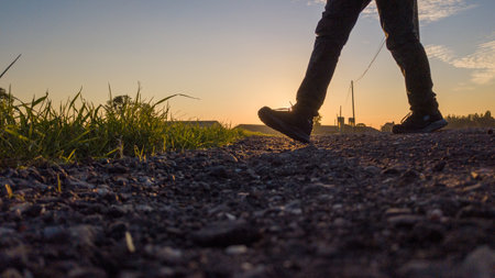 This image showcases a low-angle view of an individuals legs as they walk on a rural path at sunset. The golden light of the setting sun creates striking silhouettes and elongated shadows, highlighting the textures of the grass and gravel. Walking at Sunset on a Rural Path. High quality photoの写真素材