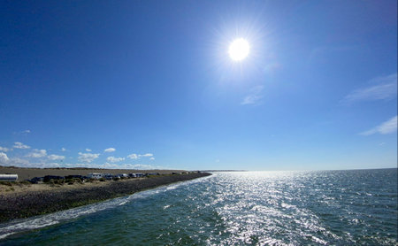 This vibrant image captures a bright sunny day at the seaside, featuring a glistening sea reflecting the sunlight, a clear blue sky, and a stony shoreline that leads to a distant horizon. The suns brilliance adds warmth to the scene. Sunny Seaside with Sparkling Waters and Clear Blue Sky. High quality photoの写真素材